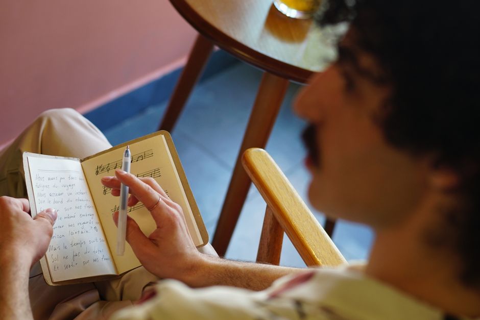 A man with a mustache writes in a notebook containing musical notations, seen from behind.